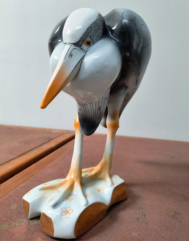 Fehér és szürke porcelán madárfigura lehajtott fejjel, narancsszínű csőrrel és lábakkal, virágmintás talapzaton.