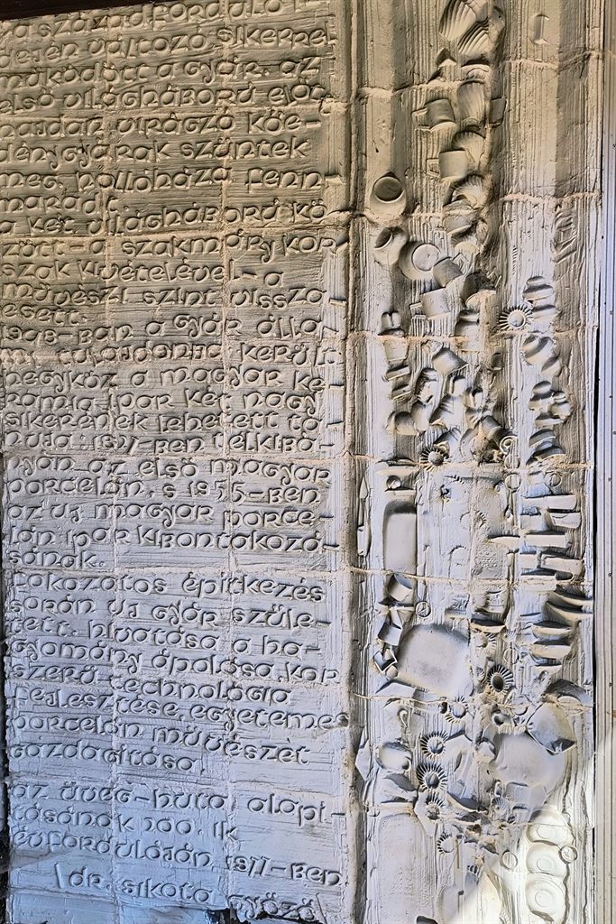 Porcelánfal domború felülete: bal oldalon bekarcolt kézírásos szöveg mezője, jobbra porcelánelemekből rakott plasztikus sáv.