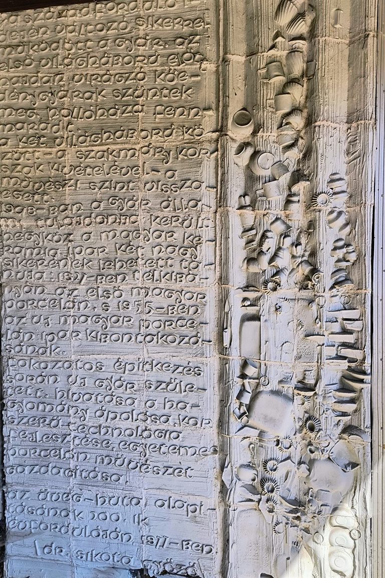 Porcelánfal domború felülete: bal oldalon bekarcolt kézírásos szöveg mezője, jobbra porcelánelemekből rakott plasztikus sáv.