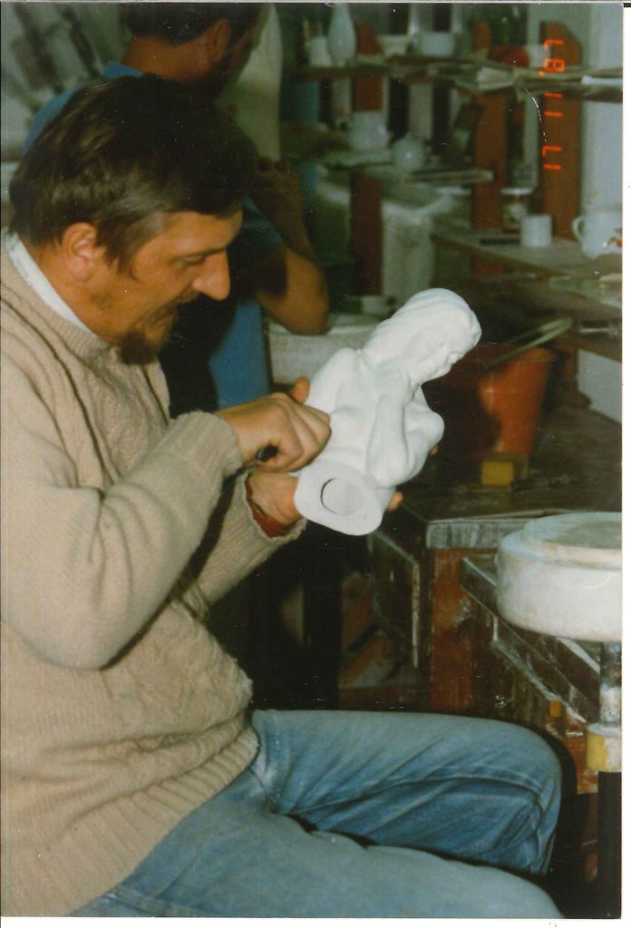 Férfi a munkapadnál fehér porcelánfigurát formáz, mögötte gipszformák és szerszámok.