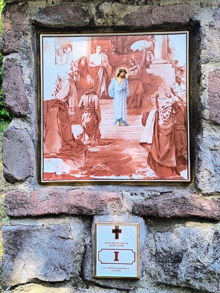 Jézus fehér ruhában áll a lépcsőn, körülötte a főpapok és a kiáltozó tömeg alakjai. Négyzetes porcelánkép aranyszínű keretben, terméskő pillérbe falazva; alatta kétnyelvű tábla a stáció számával.