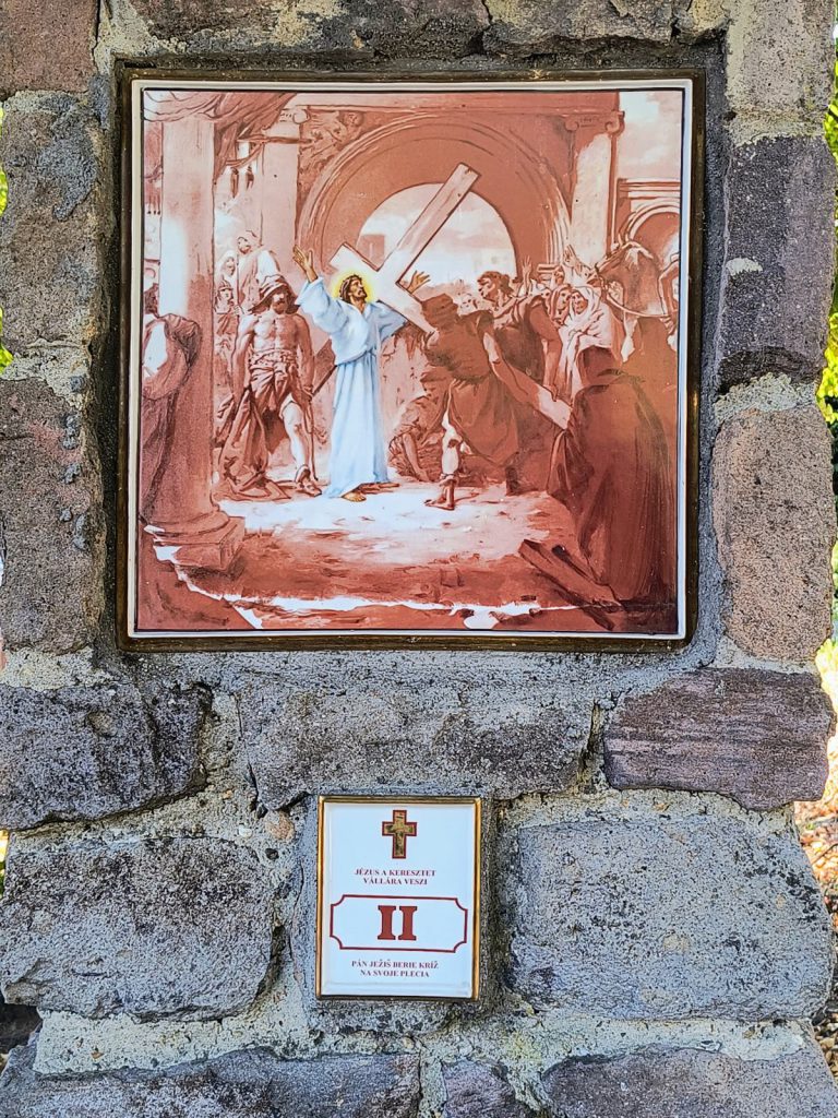 Jézus a vállára emeli a keresztet egy boltív előtt, körülötte katonák és térdelő alakok. Négyzetes porcelánkép aranyszínű keretben, terméskő pillérbe falazva; alatta kétnyelvű tábla a stáció számával.