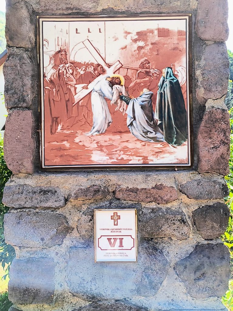 Térdelő asszony kendőt nyújt a keresztet vivő Jézusnak, a háttérben lovas katona és a városfal. Négyzetes porcelánkép aranyszínű keretben, terméskő pillérbe falazva; alatta kétnyelvű tábla a stáció számával.