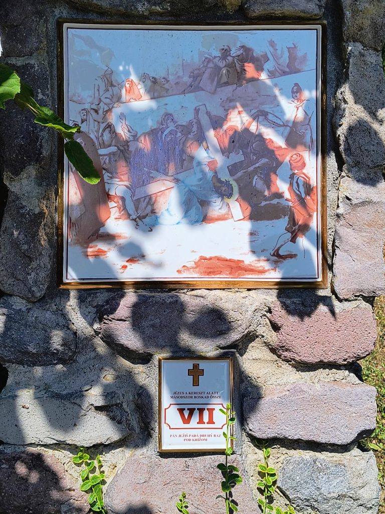 Jézus másodszor esik el a kereszt alatt; a porcelánkép felületén lombok árnyéka fut végig. Négyzetes porcelánkép aranyszínű keretben, terméskő pillérbe falazva; alatta kétnyelvű tábla a stáció számával.