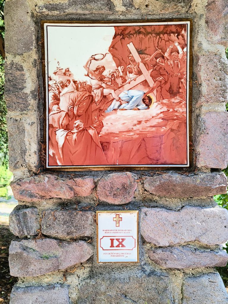Jézus harmadszor esik el, a kereszt keresztben fekszik fölötte, körülötte kiáltozó alakok. Négyzetes porcelánkép aranyszínű keretben, terméskő pillérbe falazva; alatta kétnyelvű tábla a stáció számával.