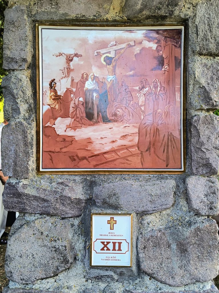 A három kereszt a Golgotán, a középsőn Jézus; alattuk Mária, János és a gyászolók. Négyzetes porcelánkép aranyszínű keretben, terméskő pillérbe falazva; alatta kétnyelvű tábla a stáció számával.