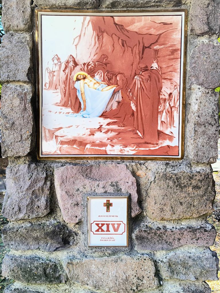 Jézus testét fehér lepelben a sziklasírba fektetik, körülötte gyászoló alakok. Négyzetes porcelánkép aranyszínű keretben, terméskő pillérbe falazva; alatta kétnyelvű tábla a stáció számával.