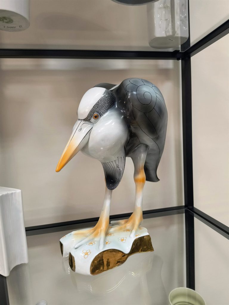 Szürke-fehér porcelán gémfigura hosszú, narancssárga csőrrel, világos talapzaton.
