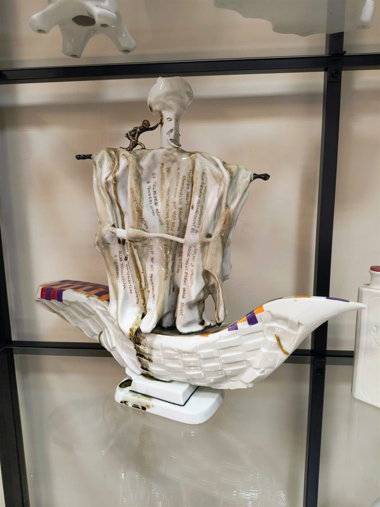 Szindbád a hajós porcelánszobra vitrinben: fehér, vitorlaszerű test bekarcolt szöveggel, tetején kis figurával.
