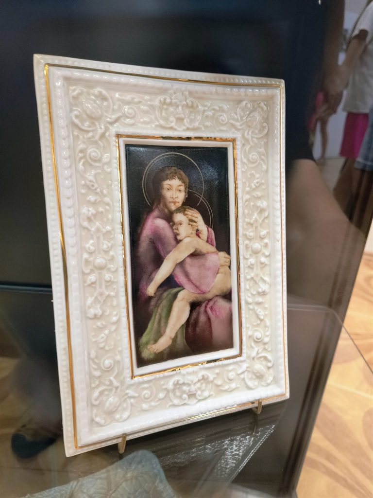Madonnát gyermekkel ábrázoló festett kép, plasztikus, fehér, virágmintás kereben.