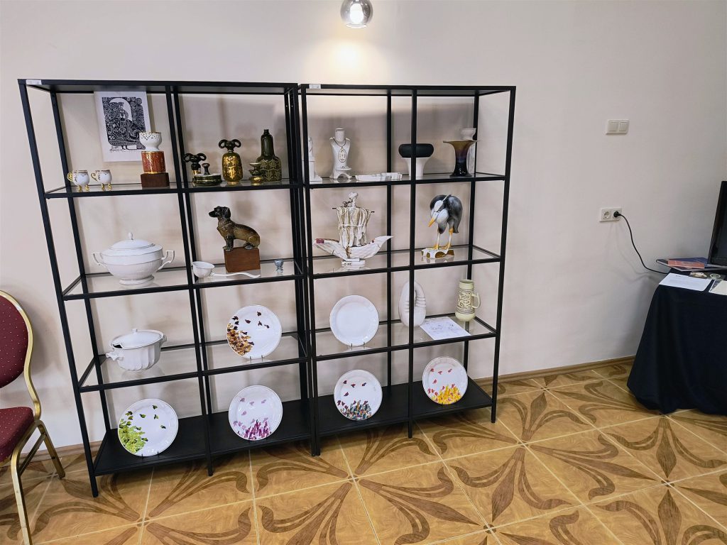Fekete fémvázas, üvegpolcos állvány porcelántányérokkal, figurákkal és vázákkal, parkettás teremben.