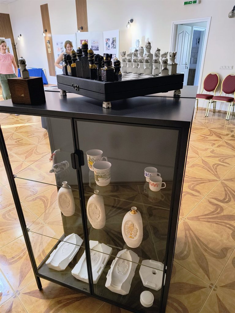 Fekete-fehér porcelán sakk-készlet sakktáblán, üvegvitrin tetején; a vitrinben csészék és porcelán plakettek.