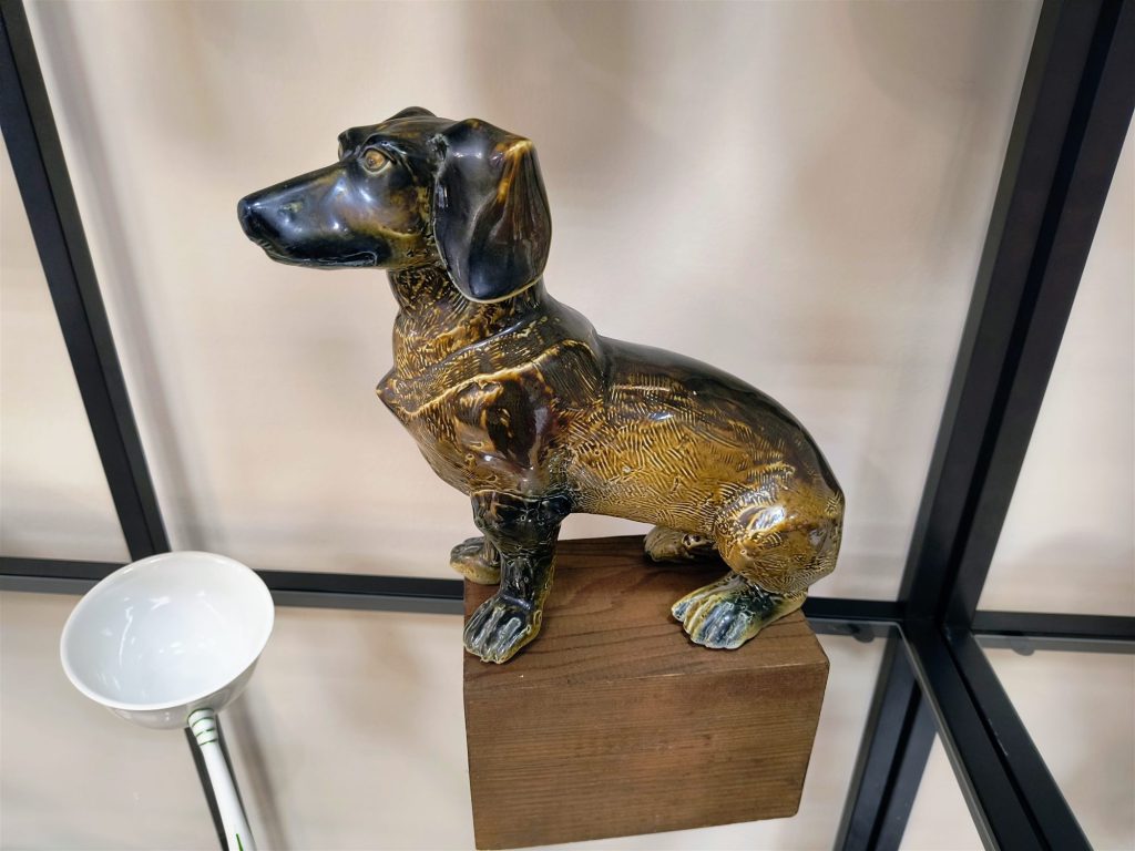 Barnás-arany mázas, ülő porcelán tacskófigura fatalapzaton.