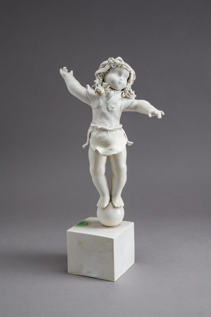 Fehér porcelán akrobatafigura világos talapzaton, szürke háttér előtt.