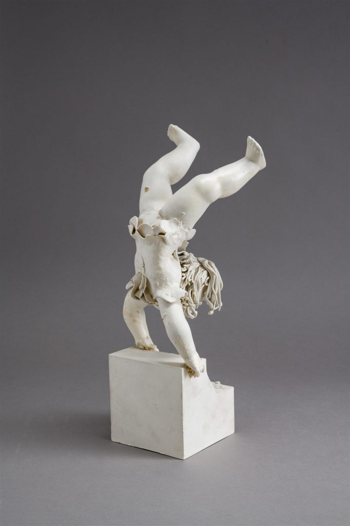 Fehér porcelán akrobatafigura világos talapzaton, szürke háttér előtt.