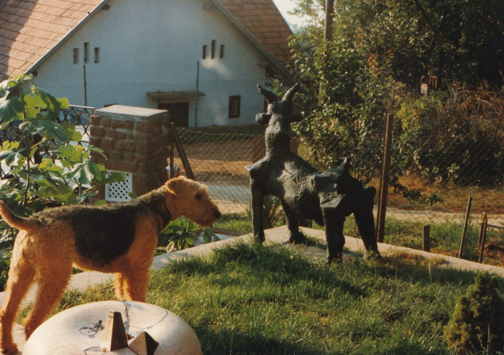 Kecske, kerámia plasztika egy kertben, előtte egy kutya